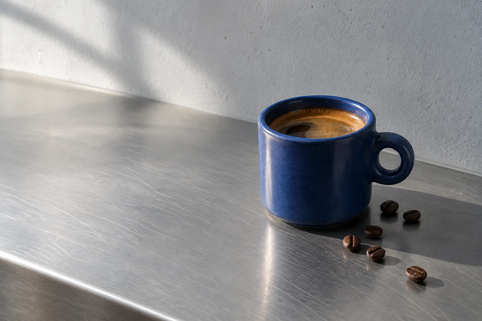 A cobalt ceramic cup of coffee on a brushed steel counter in morning light.